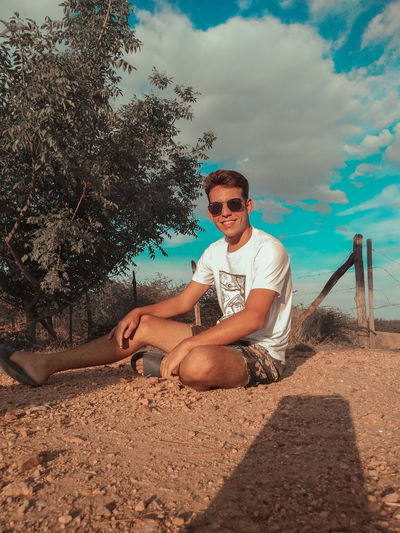 Portrait of young man wearing sunglasses sitting on land against sky