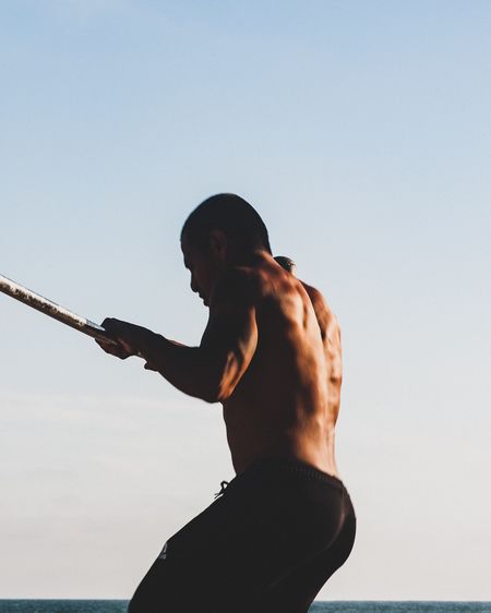 Side view of shirtless man exercising by sea against clear sky