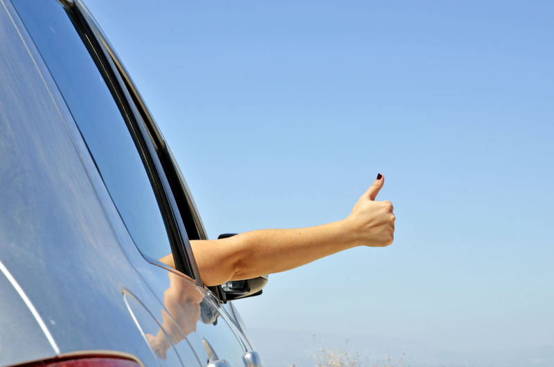 Cropped hand of woman gesturing in car window against blue sky
