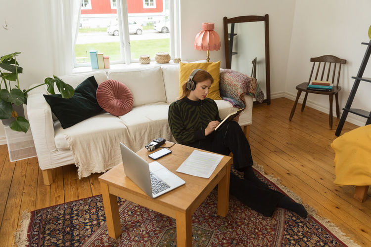 High angle of thoughtful female radio host writing in notebook while sitting on floor at home and recording podcast