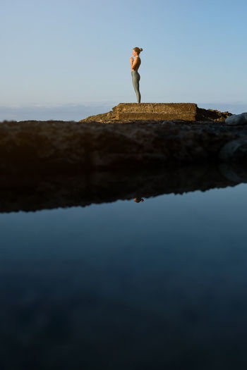 Full body side view of distant female in sportswear standing in mountain pose with prayer hands and meditating during yoga practice on rocky pier at seaside