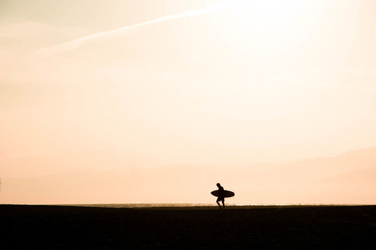 Silhouette man carrying skateboard walking on land against sky during sunset