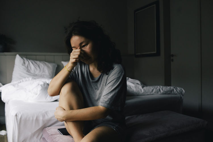 Sad woman crying while sitting on bed end bench in bedroom at home