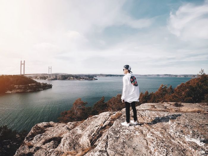 Side view of young man standing on rocky beach against cloudy sky