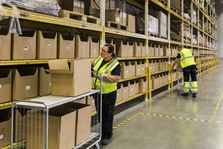 Full length of female worker pushing cart with cardboard boxes by rack against coworker at warehouse