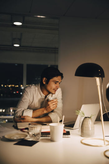 Male entrepreneur working late while using laptop sitting with coffee cup at desk in office