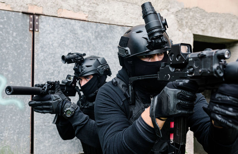 Full body of unrecognizable special forces fighters in battle gear and protective helmets aiming with modern rifles near old building walls