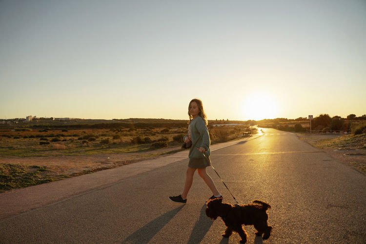Rear view of girl crossing road with dog against clear sky during sunset