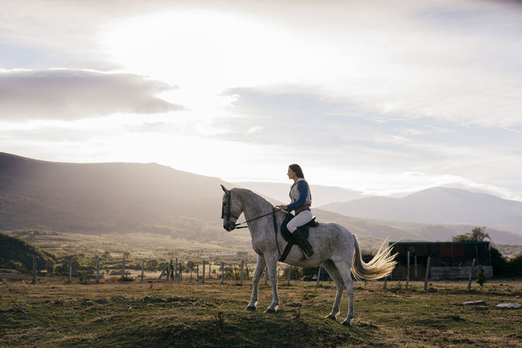 Side view of woman in hat riding a beautiful white horse on background of rural landscape under bright sky