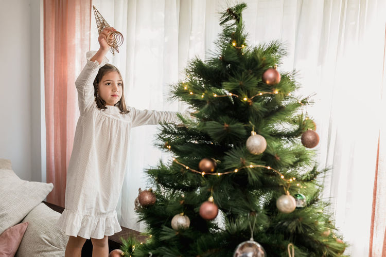 Serious girl standing on sofa and decorating coniferous christmas tree with star for celebrating holiday at home