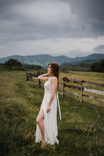 Full length of young woman standing on grassy field against cloudy sky