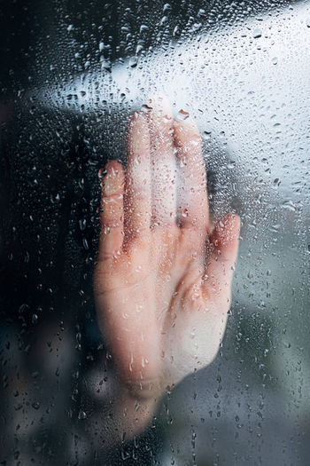Unrecognizable desperate person standing at window and touching wet steamy glass while staying in self isolation at home during coronavirus pandemic