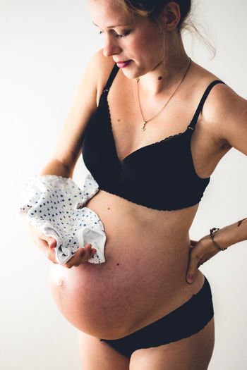 Pregnant woman in lingerie holding baby clothing against white background