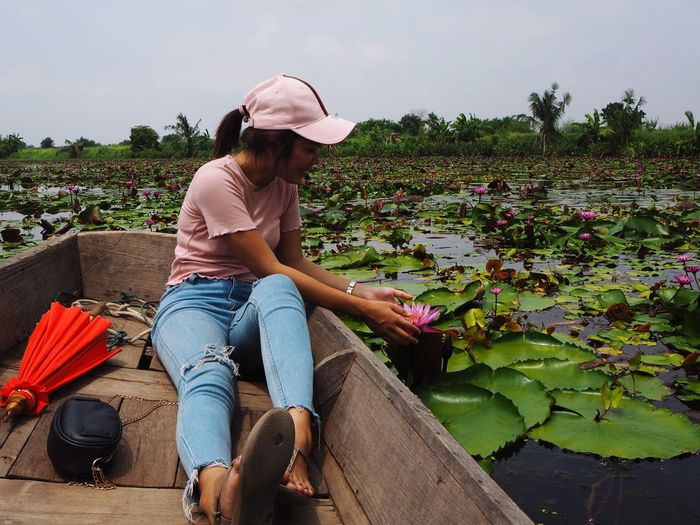Full length of young woman touching lotus water lily while sitting in boat against sky