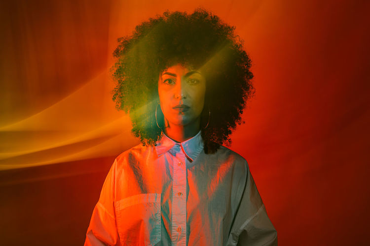 Unemotional young ethnic female model with dark afro hair in white shirt standing against red background and looking at camera