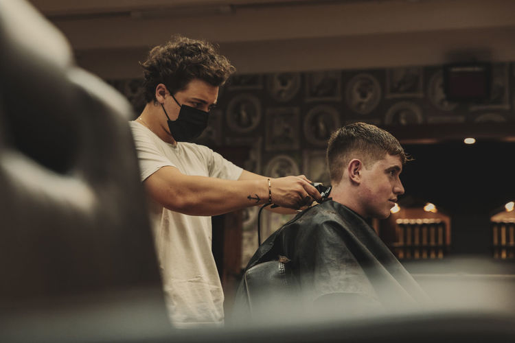 Male barber wearing face mask cutting customer's hair in salon during epidemic