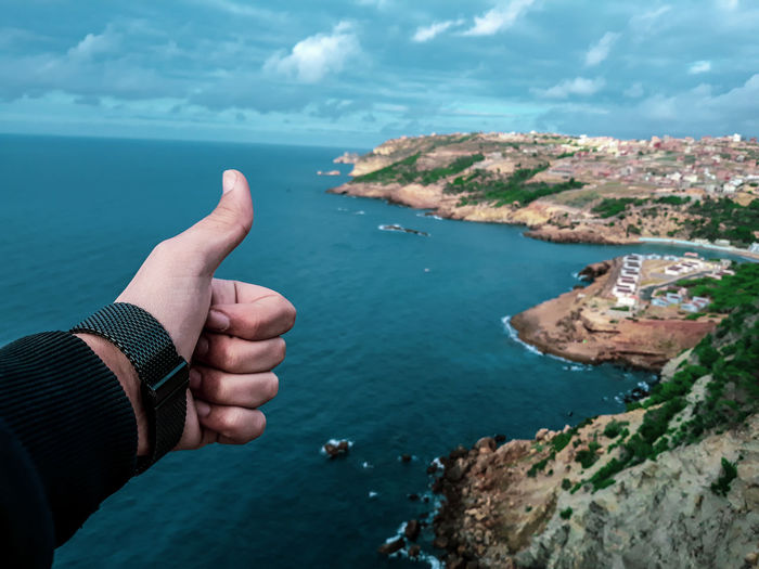 Cropped hand of man gesturing thumbs up against seascape