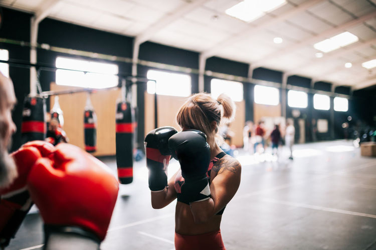 Young sportswoman athlete in boxing gloves training fighting with professional unrecognizable male coach in modern gym