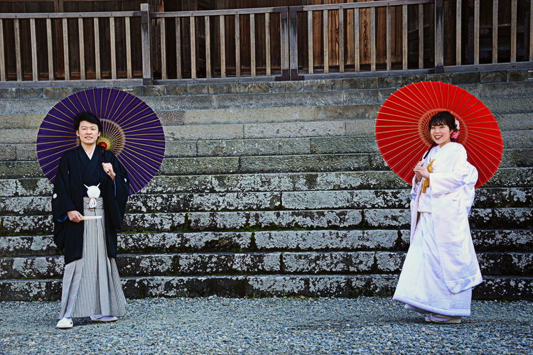 Couple wearing traditional clothing holding umbrellas while standing against wall