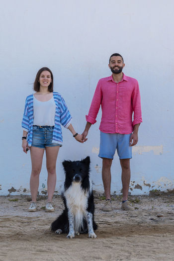 Happy couple standing with dog against wall while holding hands