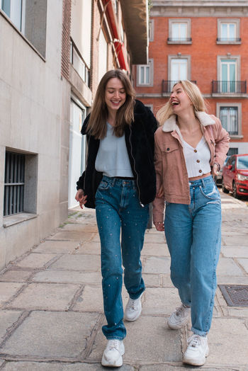 Young stylish female friends walking along street in madrid and laughing while spending weekend together and having fun