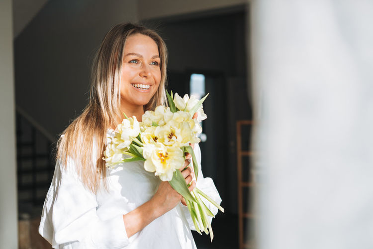 Young woman with blonde long hair in white shirt with bouquet of yellow flowers in hands at home