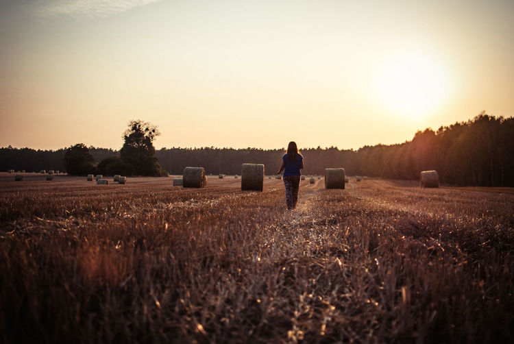 Rear view of woman walking by hay bales on field during sunset