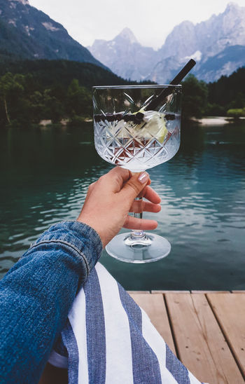 Personal perspective of woman holding glass with drink. lake, mountains in background.
