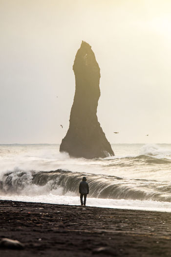 Unrecognizable back view of traveler in warm clothes and hat walking admiring dramatic landscape of volcanic coastline black sand beach with rock formations washed by foamy waves of stormy sea in iceland