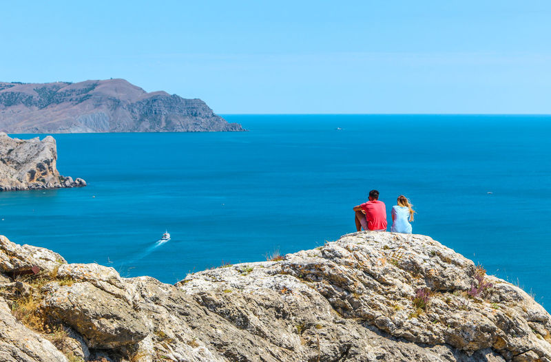 People sitting on cliff by sea against blue sky