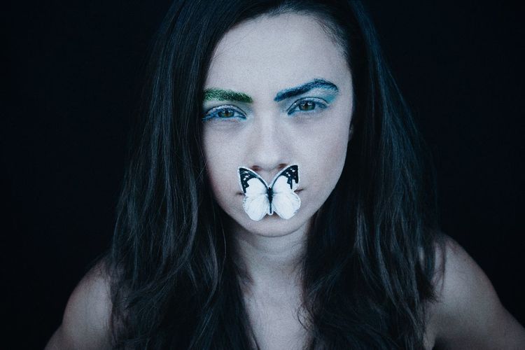 Portrait of serious woman with eye make-up and artificial butterfly on mouth against black background