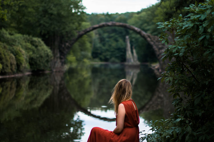Side view of woman sitting by lake with arch bridge reflection in forest