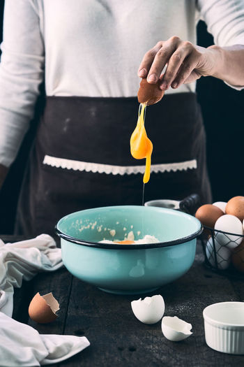 Crop faceless female cook adding egg in bowl with ingredients prepared for cooking pastry at table on black background