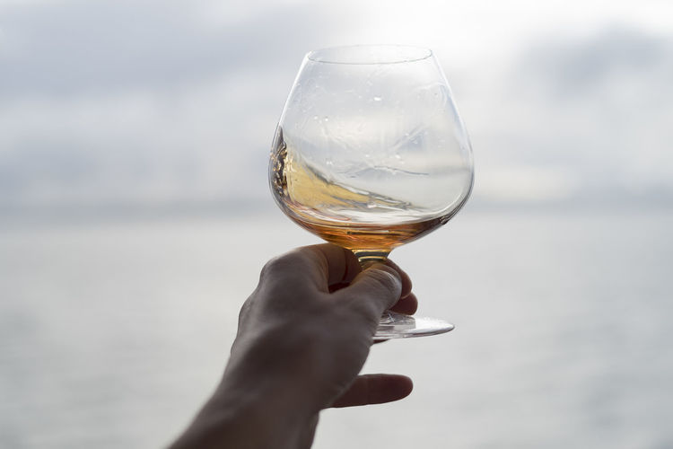 Cropped image of hand holding brandy snifter against sea