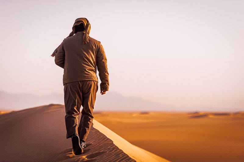 Rear view of man walking on desert against sky during sunset
