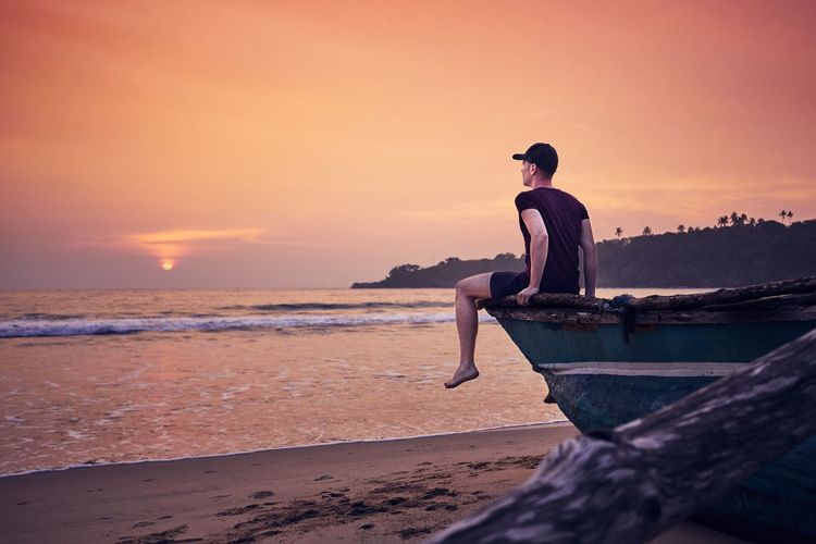 Man sitting on boat moored at beach against sky during sunset