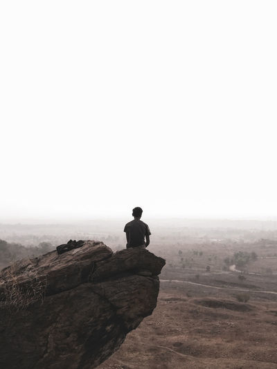 Rear view of man sitting on rock against clear sky
