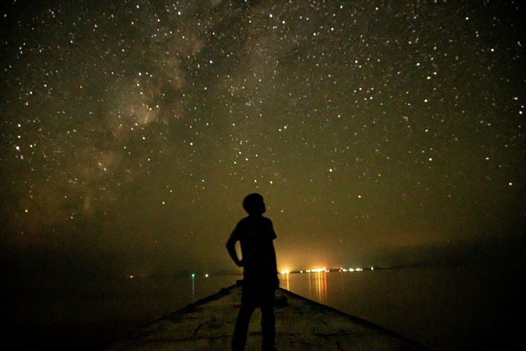 Rear view of silhouette man standing in sea against sky at night