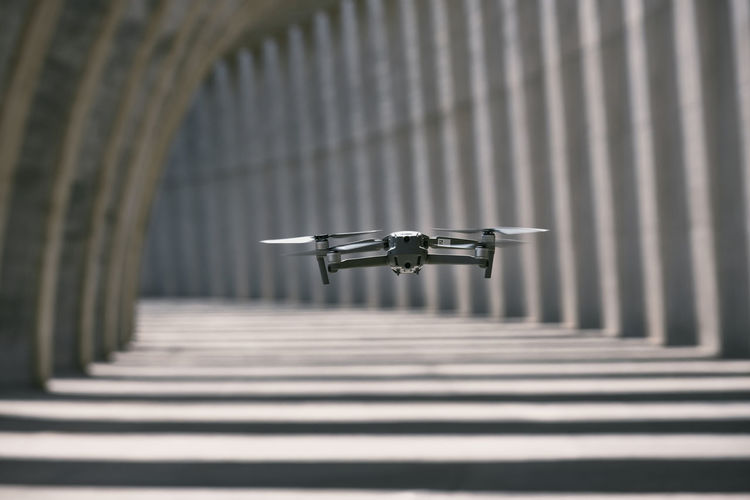 Modern drone flying in empty arched walkway with columns casting shadows in shape of lines on sunny day