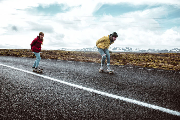 Full length of young man and woman skateboarding on road against landscape