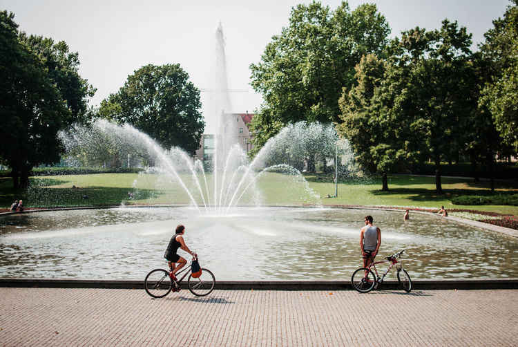 Man and woman with bicycles on footpath against fountain at park