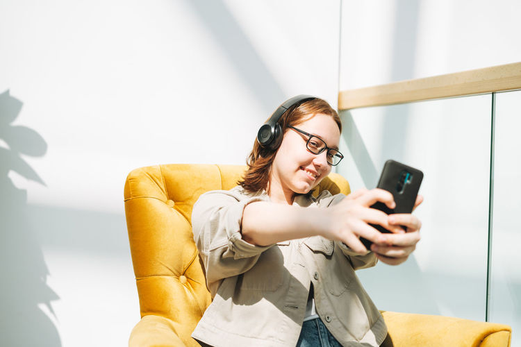 Young teenager girl student in glasses using mobile, listen music in headphones in yellow chair 