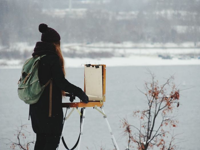 Side view of woman holding suitcase against lake during snowfall