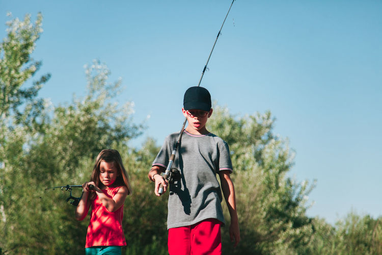 Siblings carrying fishing rods against clear blue sky