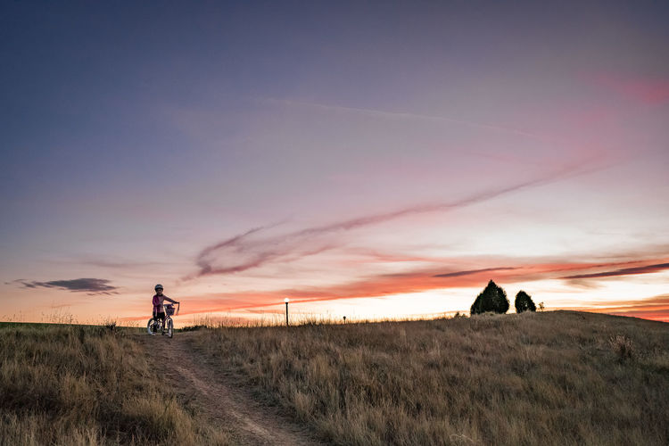 Young girl starts to bike down a dirt path on a grass hill at sunset
