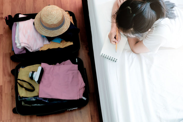 High angle view of woman writing on book with luggage in bedroom