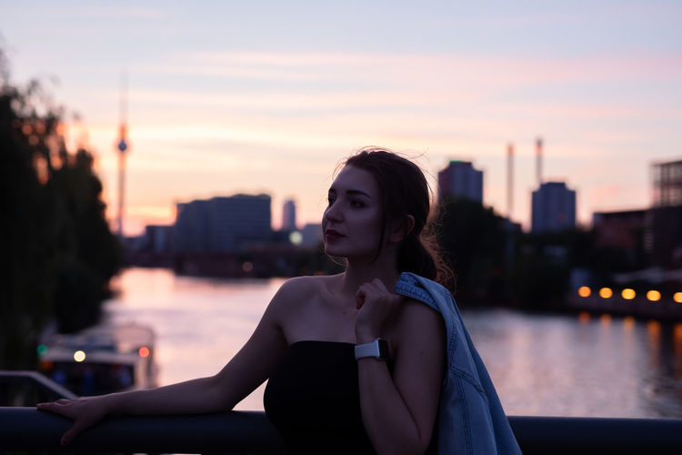 Young woman looking away while standing by railing during sunset