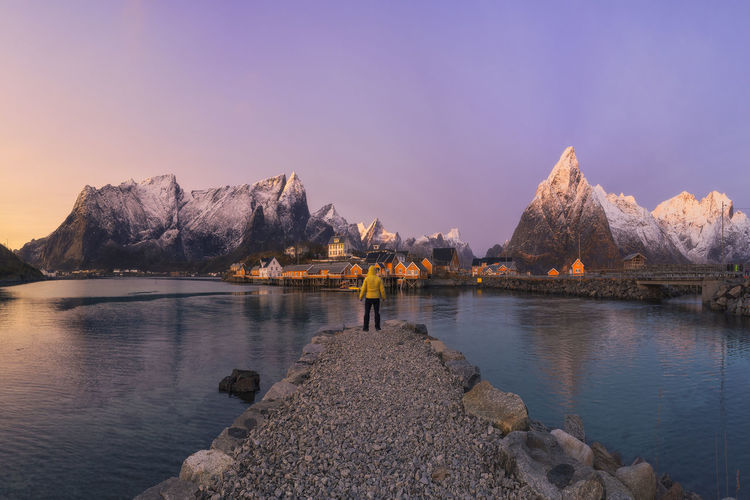 Stone quay located on calm sea water with tourist leading to reine village and snowy mountain ridge against sundown sky on lofoten islands, norway