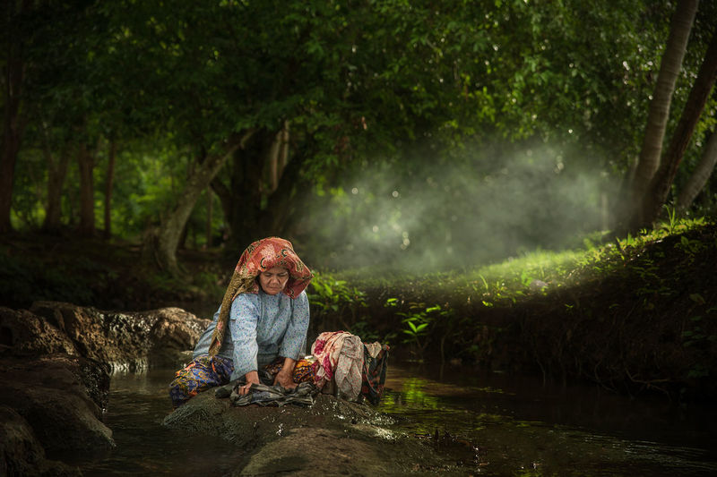 Woman washing clothes while sitting on rock in forest