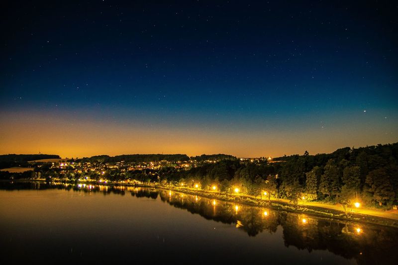 Illuminated city by lake against sky at night